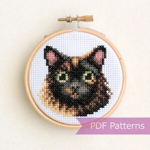 Op de afbeelding: Een kruissteekborduurwerk van een schildpadkattenkop met zwarte, bruine en oranje vacht, groene ogen en roze oren, gemonteerd in een ronde houten borduurring. Een paarse banner rechtsonder bevat de tekst PDF Patterns.