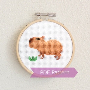 Op de afbeelding: Een kruissteekpatroon van een bruine capibara die een groen blad eet, ingelijst in een houten borduurring. De tekst "PDF Pattern" is afgedrukt op een paars lint onder de ring.