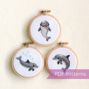 Puede incluir: Tres patrones de bordado en forma de aro que representan adorables delfines de dibujos animados. Los aros son de madera y tienen un fondo de tela blanco. Los delfines son grises y blancos y tienen diferentes poses. El texto "PDF Patterns" es visible en la parte inferior de la imagen.