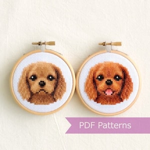 以下が含まれることがあります： 刺繍枠に入ったキャバリア・キング・チャールズ・スパニエルの顔のクロスステッチパターンが2つ。パターンはカラーで、片方のパターンでは舌を出した犬の顔、もう片方のパターンでは口を閉じた犬の顔になっています。枠の下の紫色のリボンには「PDF Patterns」という文字が印刷されています。