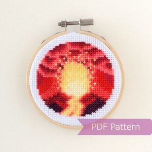 May include: A cross stitch pattern of a sunset with a yellow sun and red and orange clouds. The pattern is in a round wooden embroidery hoop. The text "PDF Pattern" is on a purple ribbon below the hoop.