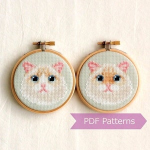 Puede incluir: Dos aros de bordado con patrones de punto de cruz de un gato blanco con marcas naranjas. El gato tiene ojos azules y una nariz rosa. Los aros son de madera y tienen un acabado natural. El texto "PDF Patterns" está impreso en una cinta morada debajo de los aros.