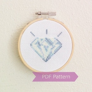 Peut inclure: Un motif de point de croix blanc représentant un diamant bleu dans un cercle de broderie en bois. Le cercle est suspendu à un mur blanc. Le texte "PDF Pattern" est imprimé sur un ruban violet sous le cercle.