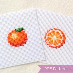 Puede incluir: Dos piezas cuadradas de tela Aida blanca con diseños de punto de cruz de naranjas. Una pieza muestra una naranja entera con una hoja verde y la otra muestra una sección transversal de una rodaja de naranja. Una banda morada tiene el texto PDF Patterns.