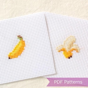 Peut inclure: Deux carrés de tissu Aida blanc avec des motifs au point de croix. L'un présente une banane jaune entière, l'autre une banane jaune partiellement pelée. Une bannière violette en bas contient le texte PDF Patterns.