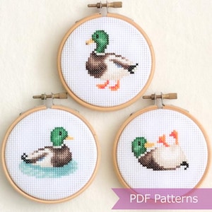 Peut inclure: Trois cerceaux à broder avec des motifs de canards en point de croix. Les cerceaux sont en bois et ont un fond en tissu blanc. Les canards sont représentés dans différentes poses, notamment en train de nager et de se tenir debout. Le texte "PDF Patterns" est visible en bas de l'image.