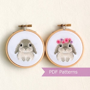 May include: Two cross-stitch patterns of a grey bunny, one with a pink flower crown. The patterns are displayed in wooden embroidery hoops. The text "PDF Patterns" is displayed below the hoops.