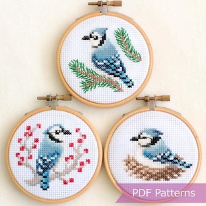 以下が含まれることがあります： 青い鳥が描かれた刺繍枠のパターンが3つあります。最初の模様は、松の枝にとまっている青い鳥です。2番目の模様は、赤い実のついた枝にとまっている青い鳥です。3番目の模様は、巣にいる青い鳥です。パターンには「PDF Patterns」とラベルが付けられています。