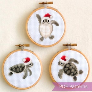 May include: Three round embroidery hoops with white fabric and cross-stitch designs of sea turtles wearing Santa hats. The hoops are made of wood and have a natural finish. The text "PDF Patterns" is printed on a purple banner below the hoops.