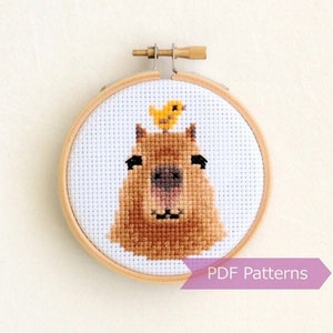 May include: A cross-stitch pattern of a capybara with a small yellow bird on its head. The pattern is framed in a wooden hoop. The text "PDF Patterns" is visible below the hoop.