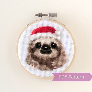 May include: A cross-stitch pattern of a sloth wearing a red Santa hat. The pattern is in a round wooden embroidery hoop. The text "PDF Pattern" is on a purple ribbon below the hoop.