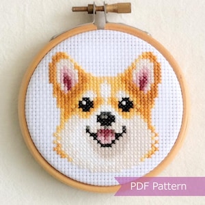 Puede incluir: Un patrón de punto de cruz que muestra la cara de un perro Corgi con pelaje dorado, hocico blanco, ojos negros y lengua rosa, exhibido en un bastidor de madera circular. El diseño está bordado sobre tela Aida blanca con el texto PDF Pattern.