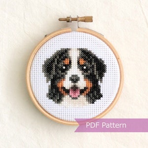 May include: Cross stitch pattern of a Bernese Mountain Dog's face in a wooden embroidery hoop. The dog has black, white, and brown fur. The pattern is available as a PDF download.