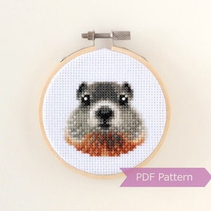 May include: A cross-stitch pattern of a grey and brown groundhog with black eyes, in a wooden embroidery hoop. The text "PDF Pattern" is on a purple banner below the hoop.