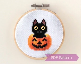 May include: Cross stitch pattern featuring a black cat sitting inside a carved orange pumpkin with a jack-o'-lantern face. The pattern is displayed in a round wooden embroidery hoop. "PDF Pattern" is printed on a purple ribbon below the hoop.