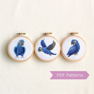 Puede incluir: Tres aros de bordado con patrones de punto de cruz de loros azules y grises. Los aros son de madera y tienen un fondo blanco. El texto "PDF Patterns" está en una cinta morada.