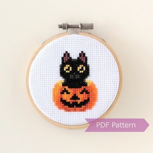 May include: Cross stitch pattern featuring a black cat sitting inside a carved orange pumpkin with a jack-o'-lantern face. The pattern is displayed in a round wooden embroidery hoop. "PDF Pattern" is printed on a purple ribbon below the hoop.