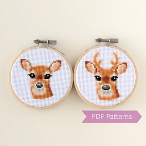 May include: Two embroidery hoops with cross-stitch patterns of deer faces. The hoop on the left features a doe with brown fur and the hoop on the right features a buck with antlers. The hoops are both white and have a natural wood finish. The text "PDF Patterns" is printed on a purple ribbon below the hoops.