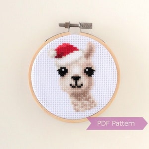 May include: A white cross-stitch pattern of a llama wearing a red Santa hat. The pattern is framed in a wooden embroidery hoop. The text "PDF Pattern" is printed on a purple ribbon.