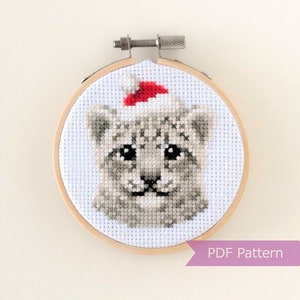 Puede incluir: Patrón de punto de cruz de un leopardo de las nieves con un gorro de Papá Noel rojo. El patrón está en un aro de bordado de madera redondo. El texto "PDF Pattern" está en una pancarta morada debajo del aro.