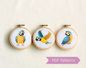 May include: Three embroidery hoops with cross-stitch patterns of blue and yellow parrots. The hoops are arranged in a row and the text "PDF Patterns" is below them.