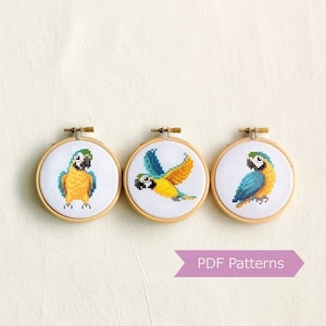 May include: Three embroidery hoops with cross-stitch patterns of blue and yellow parrots. The hoops are arranged in a row and the text "PDF Patterns" is below them.