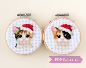May include: Two round embroidery hoops with white fabric depicting a calico cat wearing a red Santa hat. The hoops are made of wood and have a metal stand.