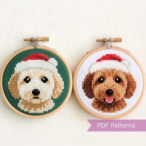 May include: Two cross-stitch hoops showing dog portraits in Santa hats. One dog is light beige on a green background, the other is brown on a white background. The hoops are in wooden frames, ideal for Christmas decorations.