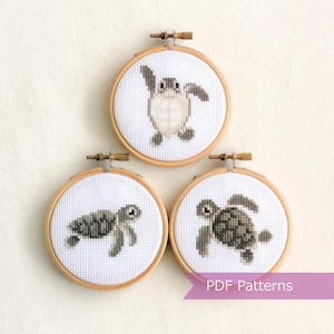 May include: Three embroidery hoops featuring cross-stitch patterns of sea turtles. The hoops are made of wood and have a white fabric background. The turtles are stitched in shades of grey and white.