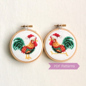 Puede incluir: Dos adornos de bordado en forma de aro que representan un gallo con un gorro de Papá Noel. El gallo es rojo, amarillo y verde. Los aros son de madera y tienen un fondo blanco. El texto "PDF Patterns" está impreso en una pancarta morada.