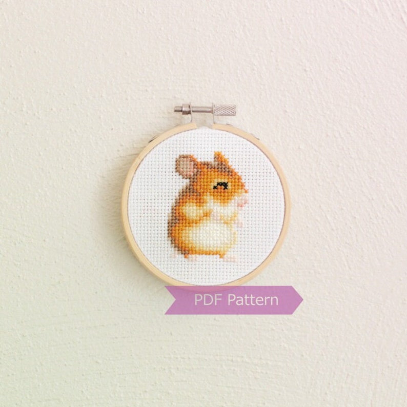 May include: A cross-stitch pattern of a cute hamster in a wooden embroidery hoop. The hamster is brown and white with a pink nose and black eyes. The text "PDF Pattern" is printed on a purple ribbon below the hoop.