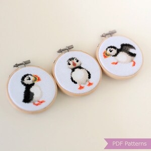 May include: Three small, round embroidery hoops with cross-stitched puffin designs. Each features a different pose of the bird, with black, white, orange, and yellow details. The hoops have wooden frames and metal tightening mechanisms.