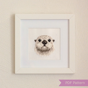 May include: A framed cross-stitch pattern of an otter's face. The otter is white with brown eyes and a black nose. The frame is white with a white mount.