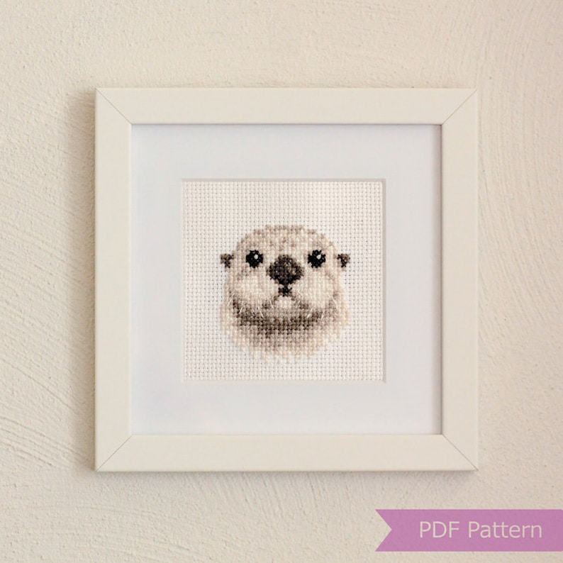 May include: A framed cross-stitch pattern of an otter's face. The otter is white with brown eyes and a black nose. The frame is white with a white mount.