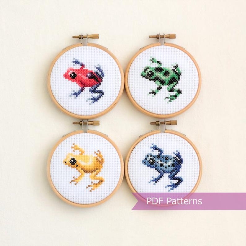 May include: Four cross-stitch embroidery hoops featuring colorful frog designs. Each frog is a different color: red, green, yellow, and blue. The hoops are set against a white background, with the text "PDF Patterns" on a banner.