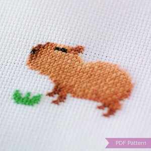Puede incluir: Patr&oacute;n de punto de cruz de un capibara marr&oacute;n comiendo una hoja verde. El patr&oacute;n est&aacute; en tela Aida blanca.