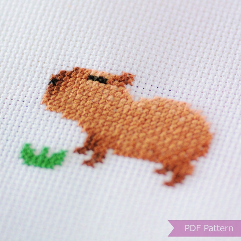 Puede incluir: Patr&oacute;n de punto de cruz de un capibara marr&oacute;n comiendo una hoja verde. El patr&oacute;n est&aacute; en tela Aida blanca.