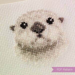 May include: Cross stitch pattern of a cute, cartoon-style otter with black eyes and a brown nose. The otter is stitched on white Aida cloth.