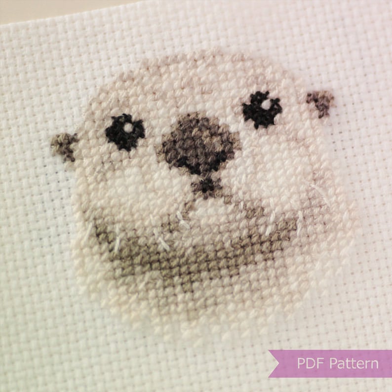 May include: Cross stitch pattern of a cute, cartoon-style otter with black eyes and a brown nose. The otter is stitched on white Aida cloth.
