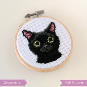 May include: Cross stitch pattern of a black cat with green eyes and pink ears. The cat is stitched on white Aida cloth and framed in a wooden hoop. The text "Green eyes" and "PDF Pattern" are visible below the image.