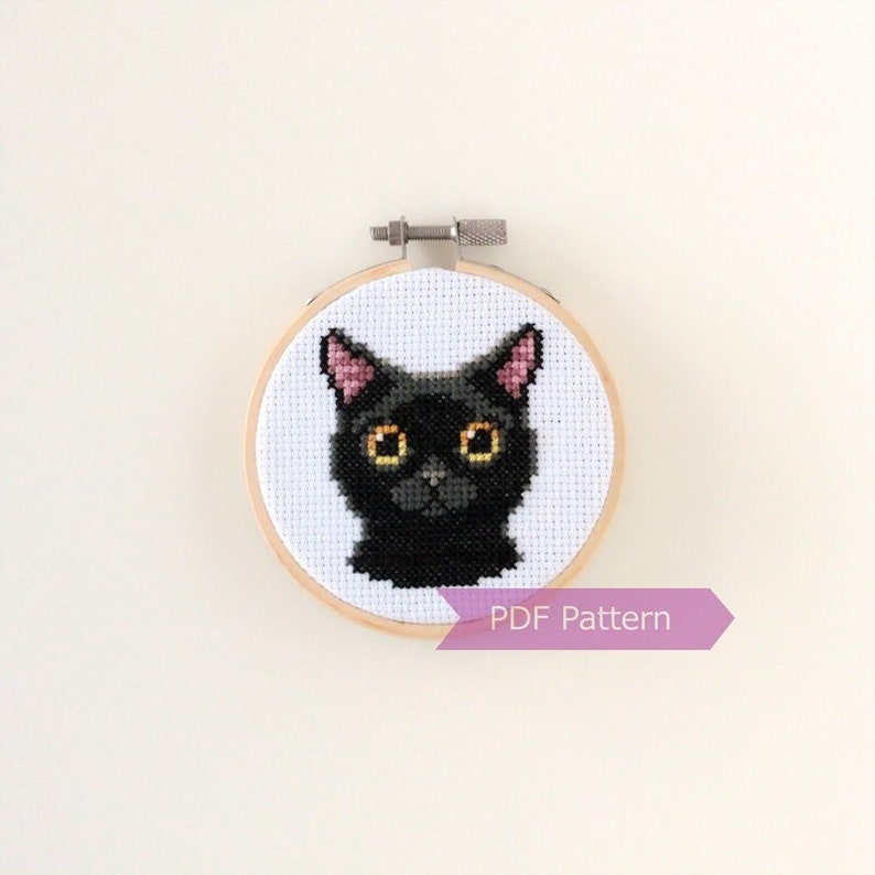 May include: A black cat cross-stitch pattern in a wooden embroidery hoop. The cat has yellow eyes and pink ears. The hoop has a white background and the text "PDF Pattern" is printed on a pink ribbon.
