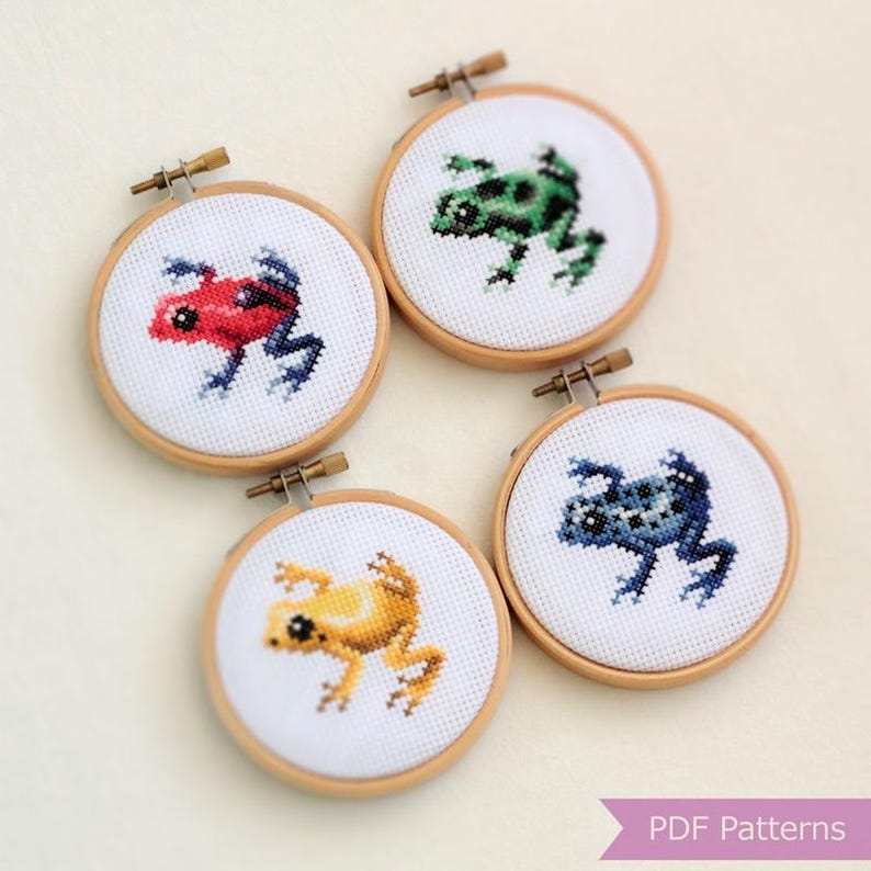 K&ouml;nnte beinhalten: Vier kleine Kreuzstich-Stickrahmen mit farbenfrohen Froschmotiven. Die Fr&ouml;sche sind rot, gr&uuml;n, gelb und blau, jeweils mit dunklen Akzenten. Die Rahmen sind aus Holz und der Stoff ist wei&szlig;. Der Text "PDF Patterns" ist enthalten.