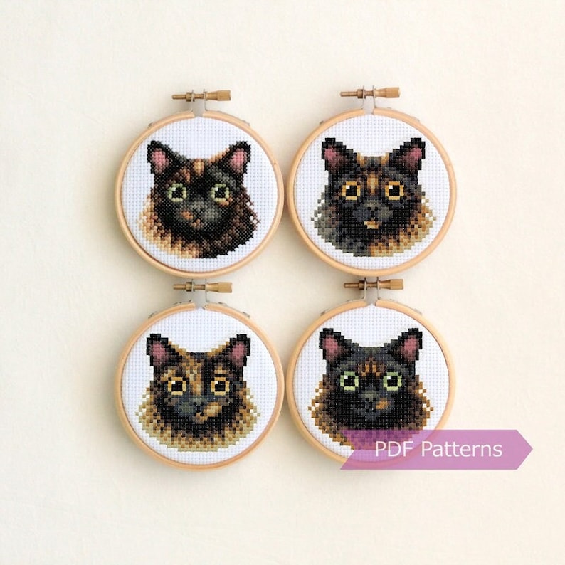May include: Four circular wooden embroidery hoops containing cross-stitch portraits of cat faces. Each hoop features a different feline with unique tortoiseshell and black fur patterns, large eyes, and pointed ears on white fabric. Text reads PDF Patterns.
