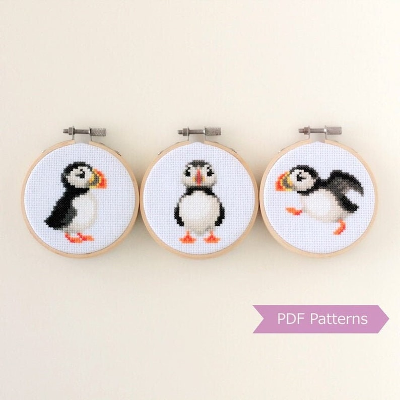 May include: Three cross-stitch puffin bird designs in wooden hoops. Each features a white background with a black, white, and orange puffin. The text "PDF Patterns" is in a purple banner below the hoops.