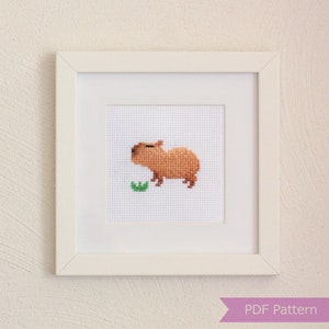 Puede incluir: Un marco blanco con una imagen de capibara marr&oacute;n en punto de cruz comiendo una hoja verde. La imagen est&aacute; titulada "PDF Pattern".