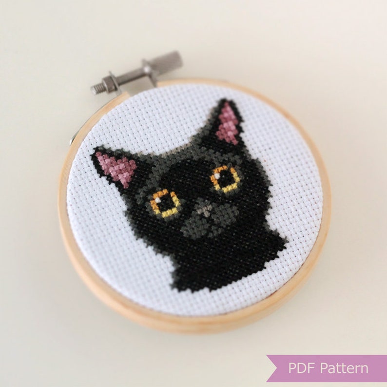 May include: Cross stitch pattern of a black cat with yellow eyes in a wooden embroidery hoop. The cat's ears are pink.