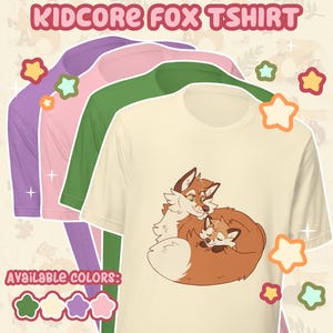 May include: Cream-colored t-shirt featuring a cartoon fox illustration, with a mother fox and baby fox cuddling. The image also shows long-sleeved shirts in green, pink, and purple. Text at the top reads "KIDCORE FOX TSHIRT".