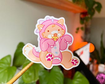 May include: A die-cut sticker featuring a cartoon fox in a pink dinosaur onesie. The fox has a pacifier, a bandage, and pink paw prints. The sticker is on a wooden stick, with green plants in the background.