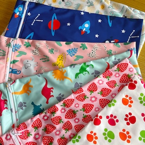 Puede incluir: Una pila de bolsas con cremallera de colores con varios diseños. Los diseños incluyen dinosaurios, fresas, temas espaciales con cohetes y planetas, y huellas de patas. Las bolsas tienen cremalleras blancas y están hechas de una tela lisa estampada.