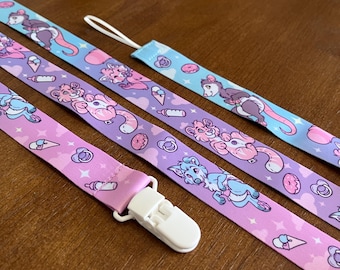 Puede incluir: Tres correas de tela con estampados de animales de dibujos animados, incluyendo zarigüeyas, gatos y perros con pañales. Las correas están decoradas con patrones de biberones, chupetes, donas y conos de helado, con un clip de plástico blanco.